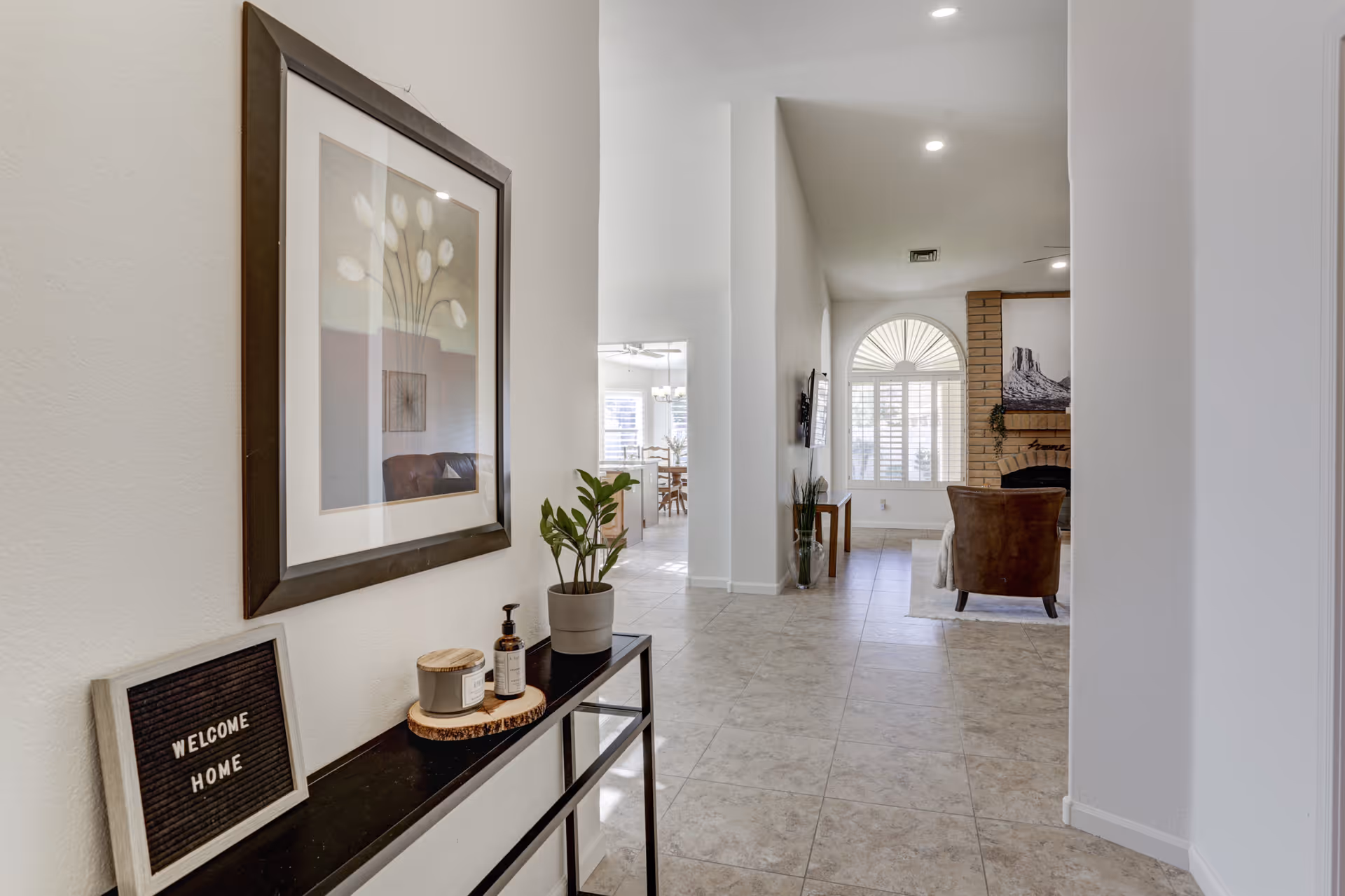 Interior view of a senior living facility hallway with a console table on the left holding a small plant, a candle, a bottle of hand soap, and a sign that reads 'WELCOME HOME'. The hallway leads to a living room area with a brown armchair, a wall-mounted TV, a large window with white shutters, and a brick fireplace with a framed picture above it. The floor is tiled and the walls are painted white.