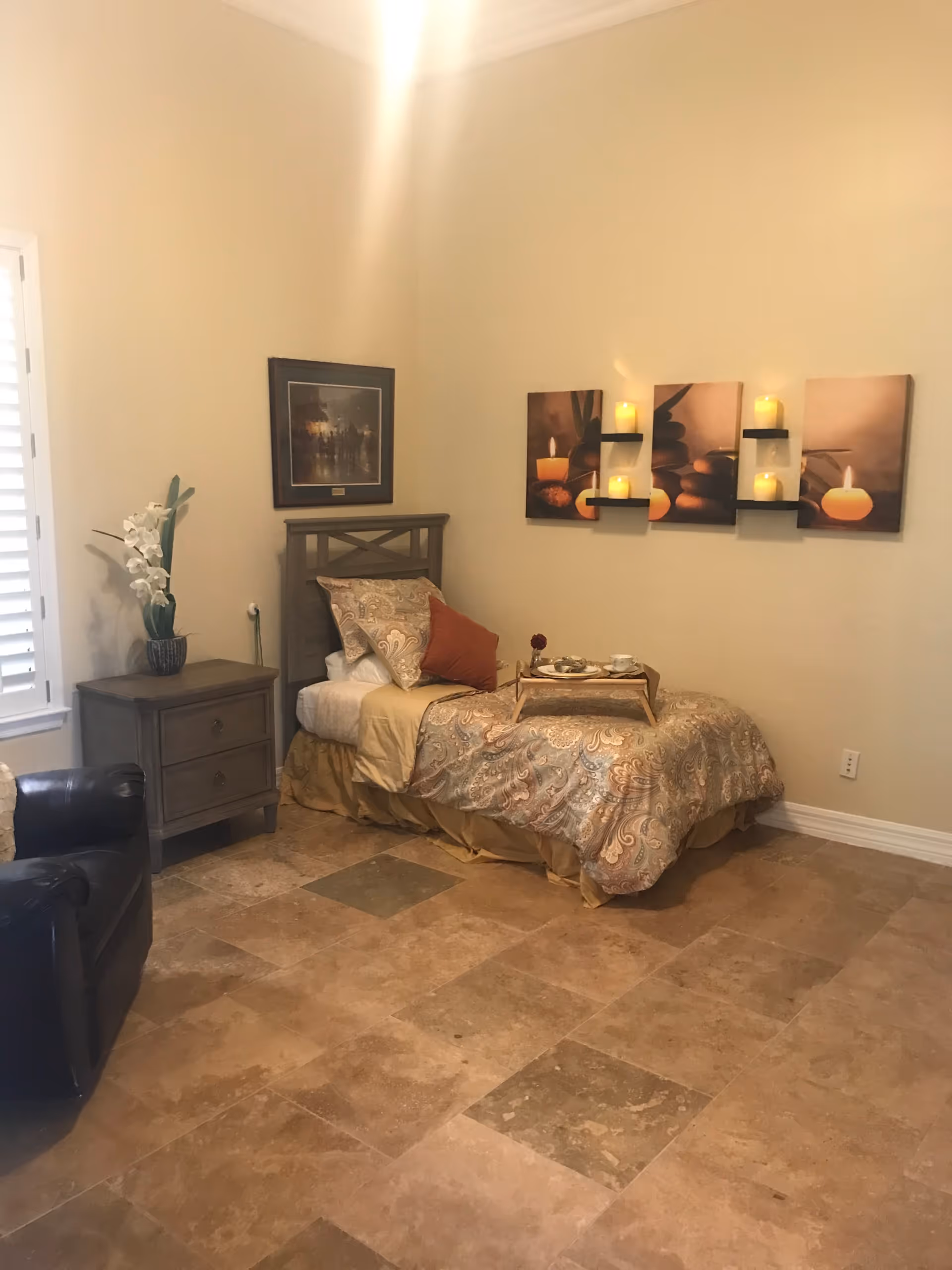 A cozy bedroom with a single bed covered in patterned bedding and pillows, a wooden nightstand with a vase of white flowers, a black leather armchair, and wall art featuring candles and stones. The floor is tiled with large beige and brown tiles.