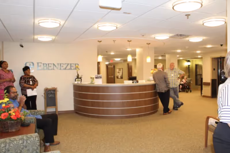 A reception area inside a senior living facility with a curved wooden front desk. Several people are standing and sitting around the area, including two women standing near a wall with the word 'EBENEZER' displayed. The space is well-lit with ceiling lights and has beige walls and carpeted floors.