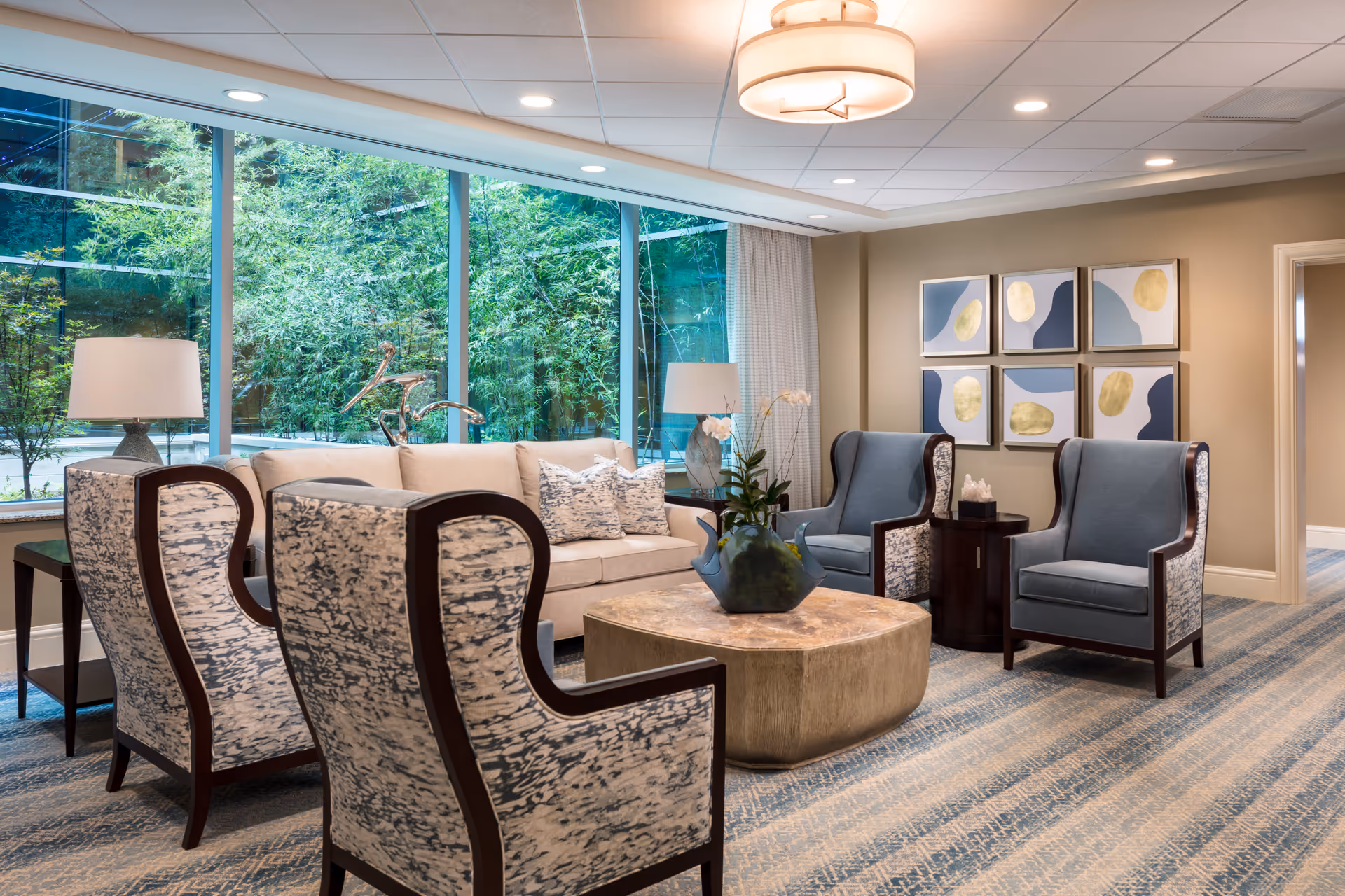 A bright and modern living room with large floor-to-ceiling windows showing green foliage outside. The room features a beige sofa with patterned pillows, four upholstered armchairs with wooden frames, a large square coffee table with a decorative vase and flowers, two table lamps on side tables, and six abstract art pieces on the wall. The carpet has a blue and beige pattern, and the ceiling has recessed lighting and a central light fixture.