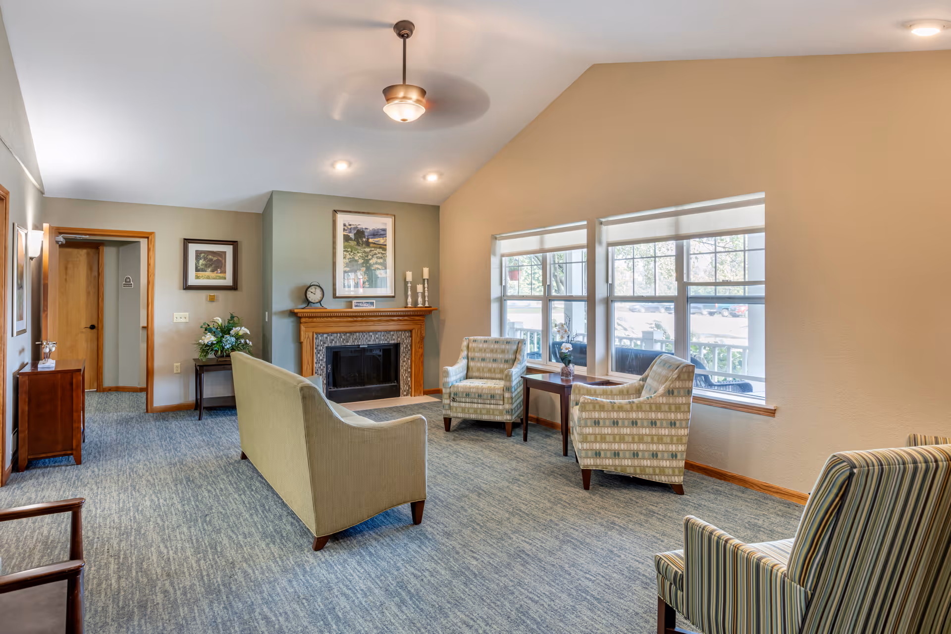 A cozy living room area with a beige sofa facing a fireplace with a wooden mantel. Two patterned armchairs and a small wooden table with a flower vase are positioned near large windows letting in natural light. The room has carpeted flooring, light-colored walls, and ceiling lights including a ceiling fan with a light fixture.