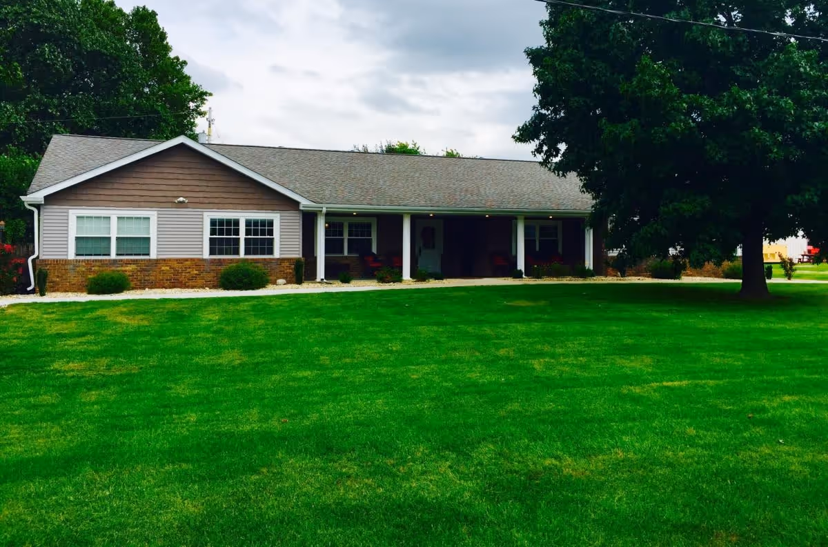 Single-story building with a covered porch, surrounded by a well-maintained green lawn and large trees under a cloudy sky.