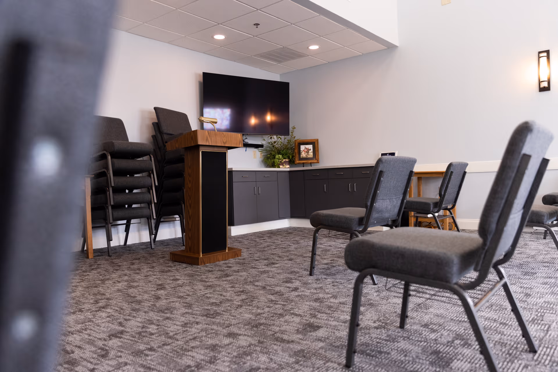 A small conference or meeting room with gray carpet, several gray padded chairs arranged facing a wooden podium with a microphone. A large flat-screen TV is mounted on the wall above gray cabinets. There is a framed picture and a plant on the cabinet. The room has white walls and ceiling lights.