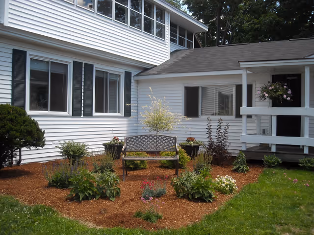 A white building with black shutters and multiple windows surrounding a small garden area. The garden has various plants and flowers with a wooden bench placed in the center. There is a small porch with a hanging flower basket on the right side of the image.