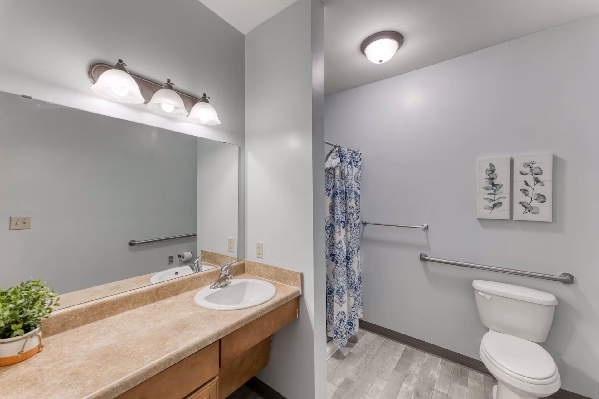 A clean and modern bathroom featuring a beige countertop with a white sink, a large mirror with three overhead lights, a toilet with a grab bar, a shower with a blue and white patterned curtain, and two botanical wall art pieces above the toilet.