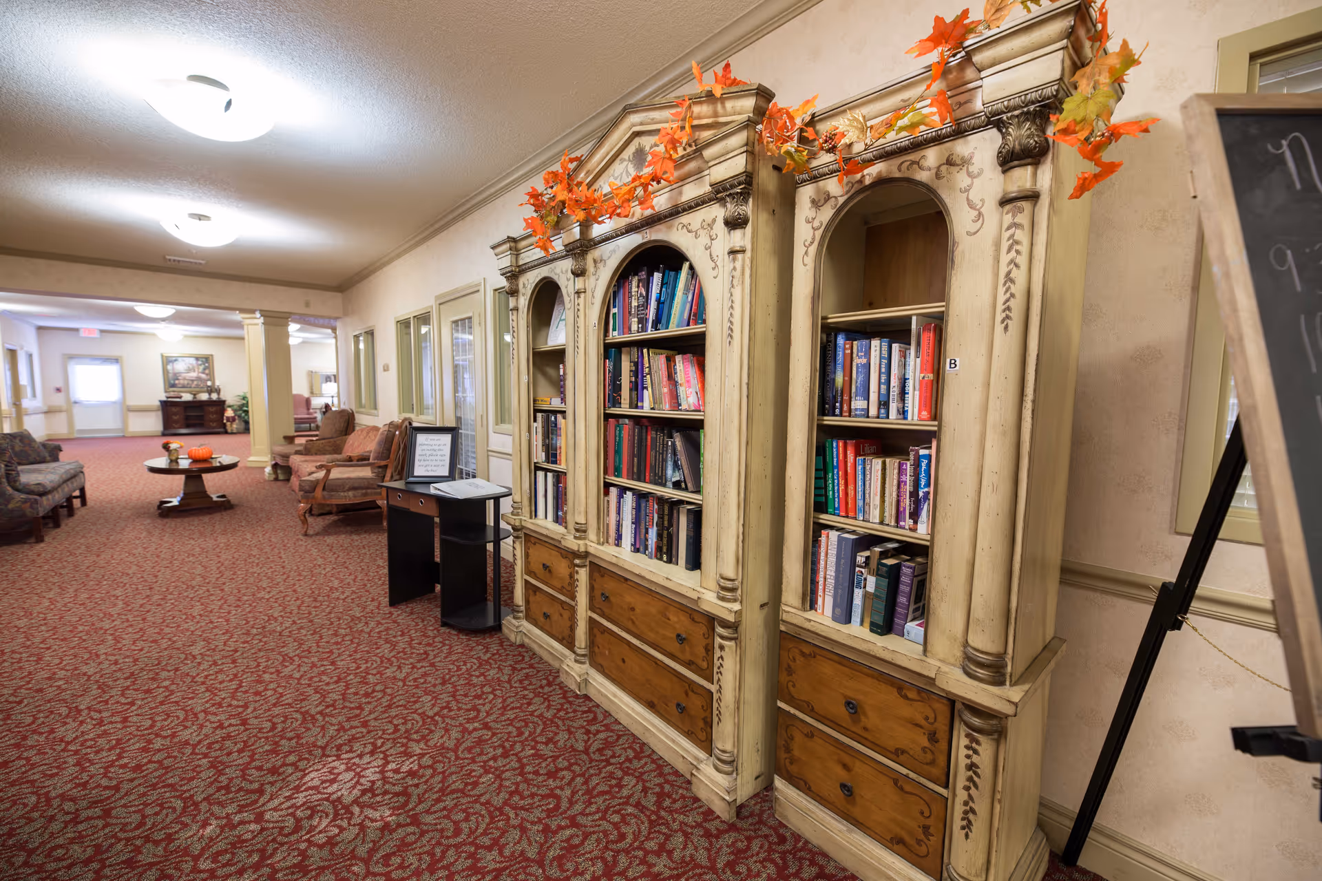 Carpeted common hallway with ornate bookshelves filled with books, seating areas and tables in a senior living facility.