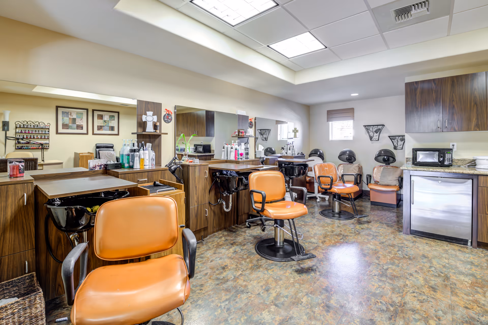Interior view of a salon area with multiple orange salon chairs, black hair washing sinks, large mirrors, wooden cabinets, and hair care products on the counters. The room has a tiled floor, a skylight ceiling, and a small window with a blind.