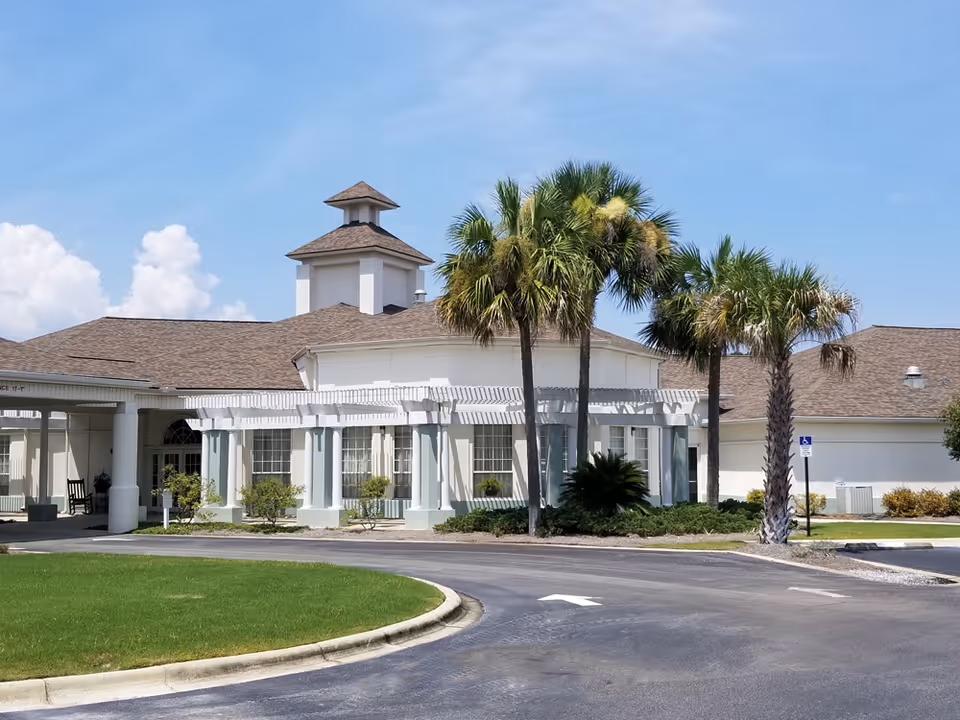 Front exterior of a retirement community building with white columns, palm trees, and a circular driveway.