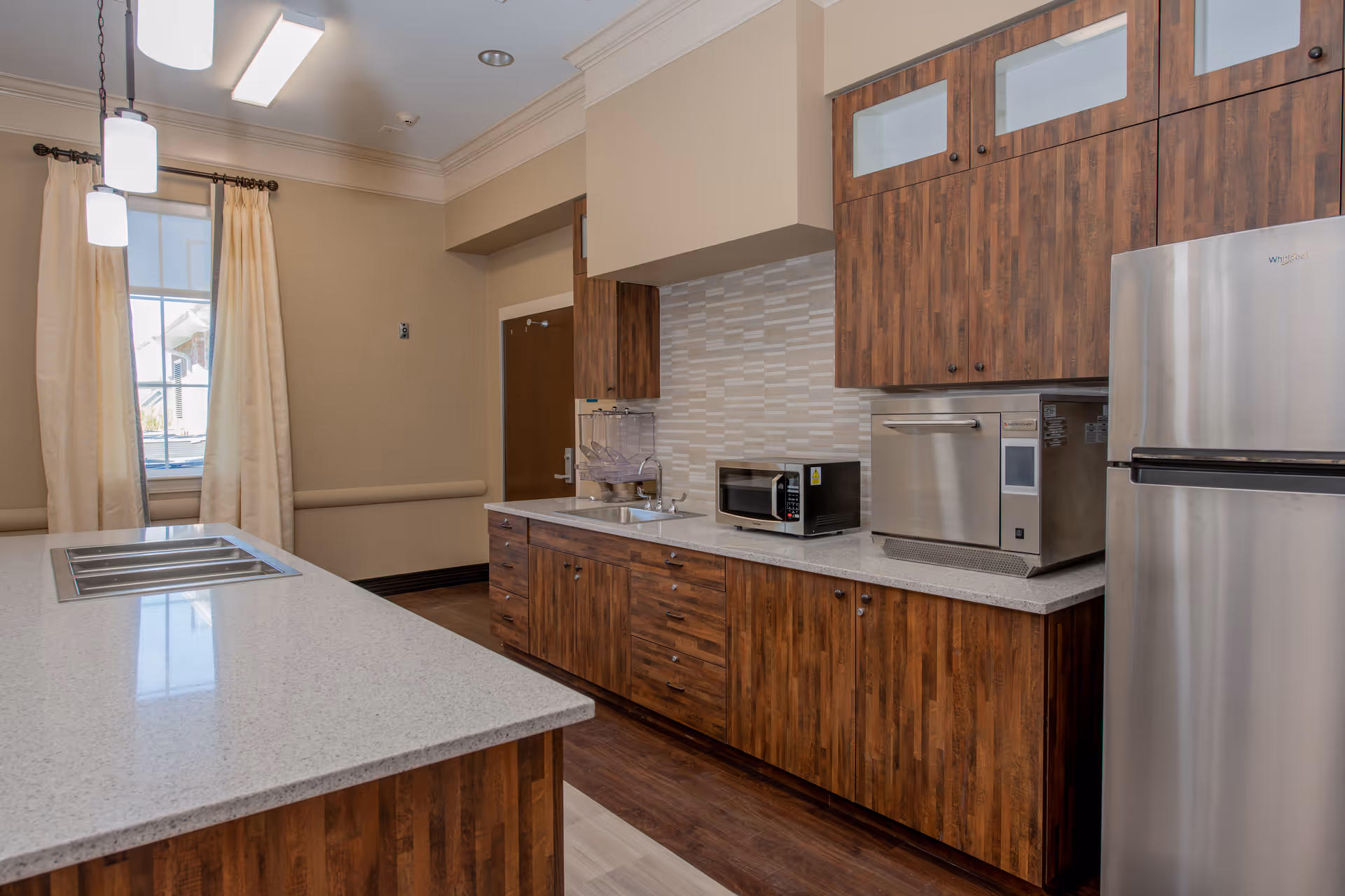 A modern kitchen with wooden cabinets, a stainless steel refrigerator, a microwave, and a countertop with a built-in sink. There is a window with cream-colored curtains letting in natural light.