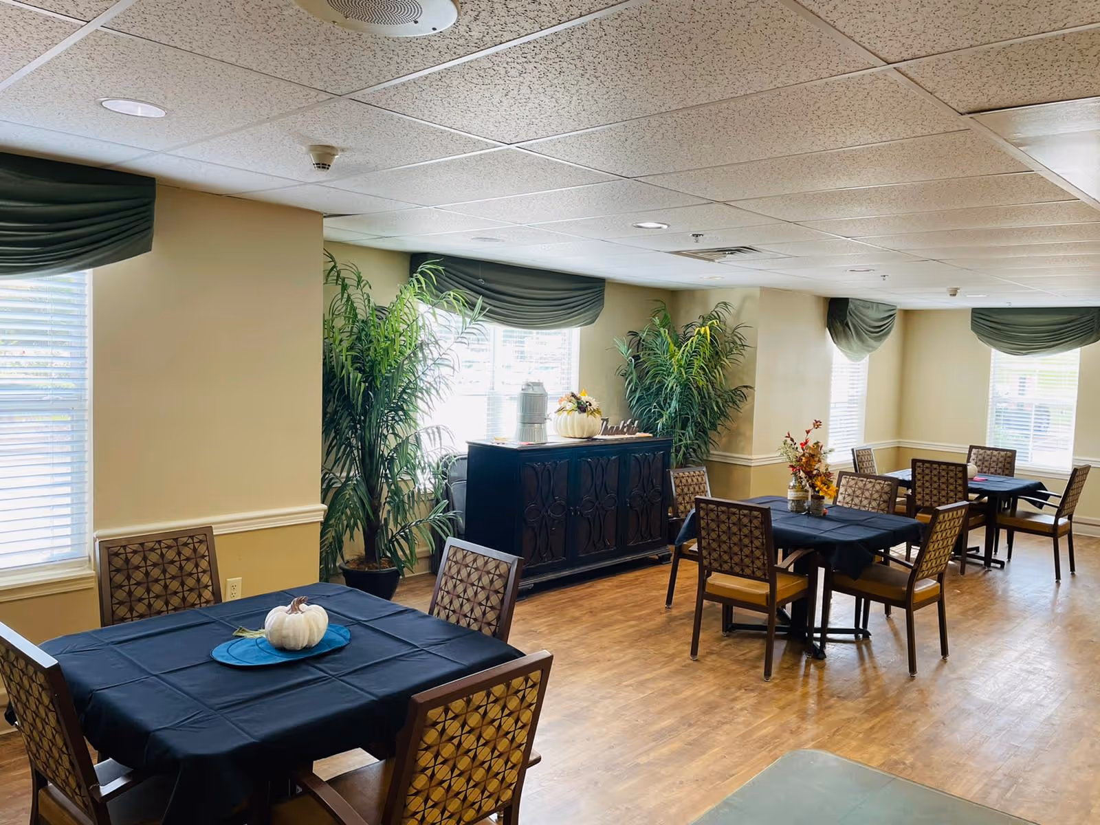 Interior view of a dining room in a senior living facility with several tables covered with black tablecloths, each surrounded by four patterned chairs. The room has large windows with green valances, potted plants, and a dark wooden sideboard with decorative items on top. The floor is wooden, and the ceiling has white tiles with recessed lighting.