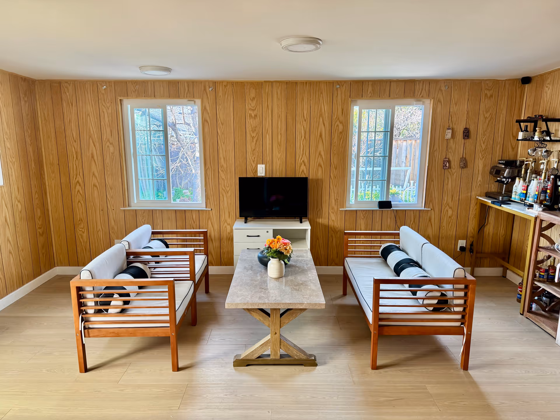 A cozy living room with two wooden sofas featuring white cushions and black-and-white cylindrical pillows facing each other across a rectangular marble coffee table with a flower vase. A small TV sits on a white cabinet against a wood-paneled wall with two windows letting in natural light. To the right, there is a small bar area with various bottles and a coffee machine.