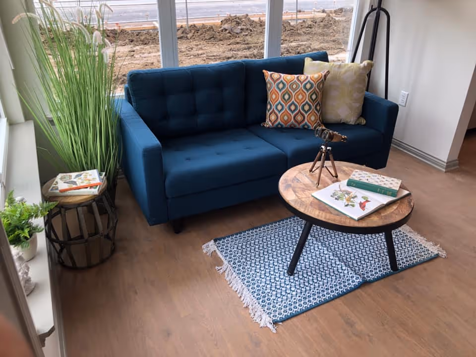 A cozy living room area with a blue tufted sofa adorned with two decorative pillows, a round wooden coffee table with books and a small telescope on it, a blue patterned rug underneath, and tall green plants placed beside the sofa near large windows showing an outdoor construction area.