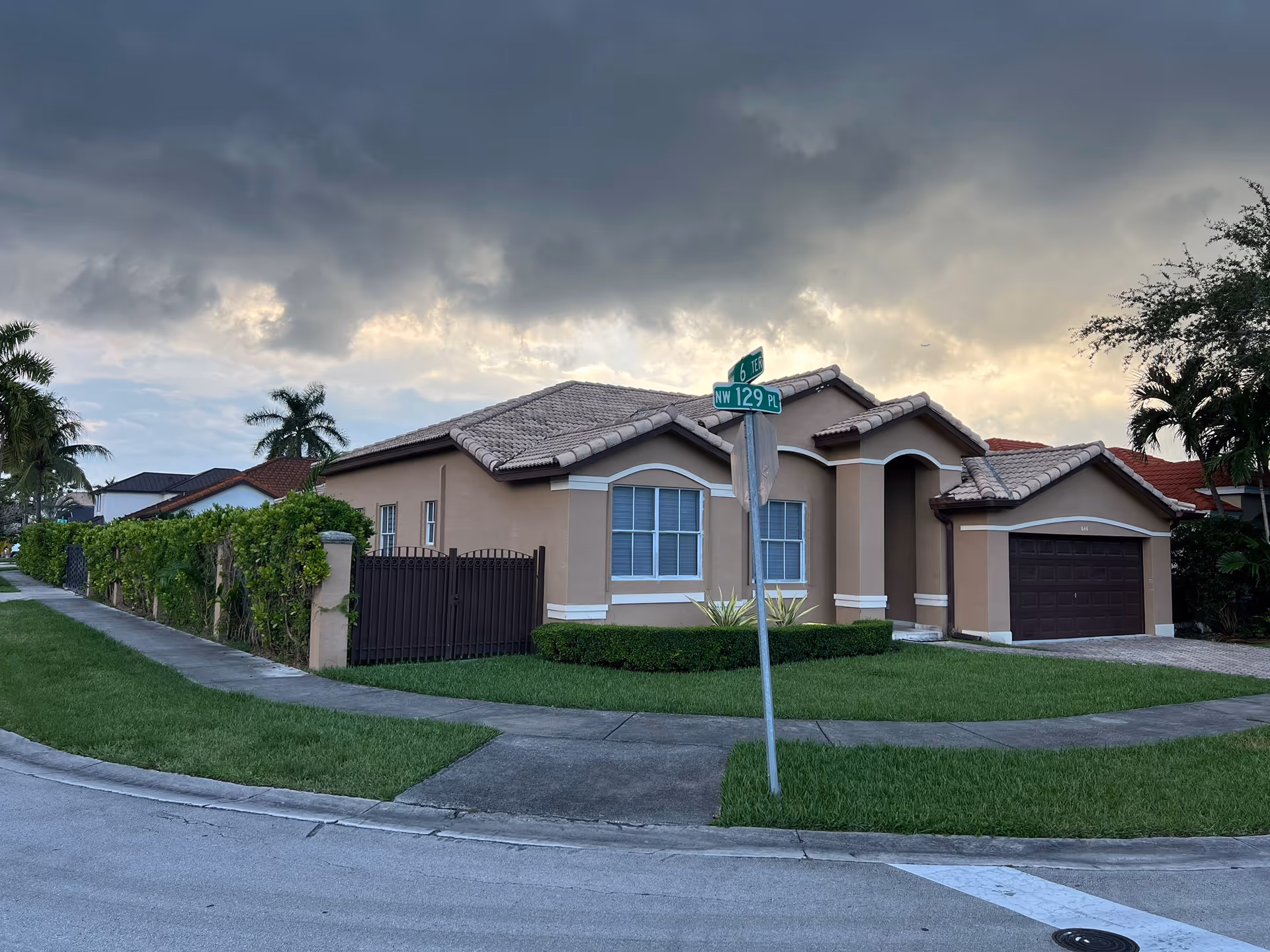 Single-story stucco house with a tiled roof and attached garage on a corner lot under a cloudy sky.