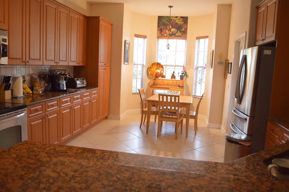 Sunny kitchen with wooden cabinetry, granite countertops, stainless appliances and a small dining table in a bay window nook.