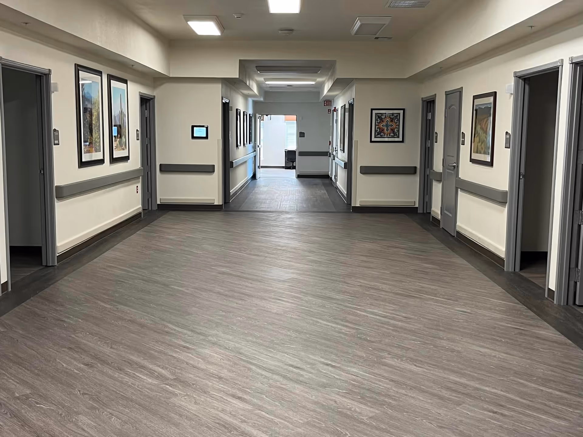 Wide interior hallway of a senior living facility with doors and framed artwork along both walls, handrails, and wood-look flooring.