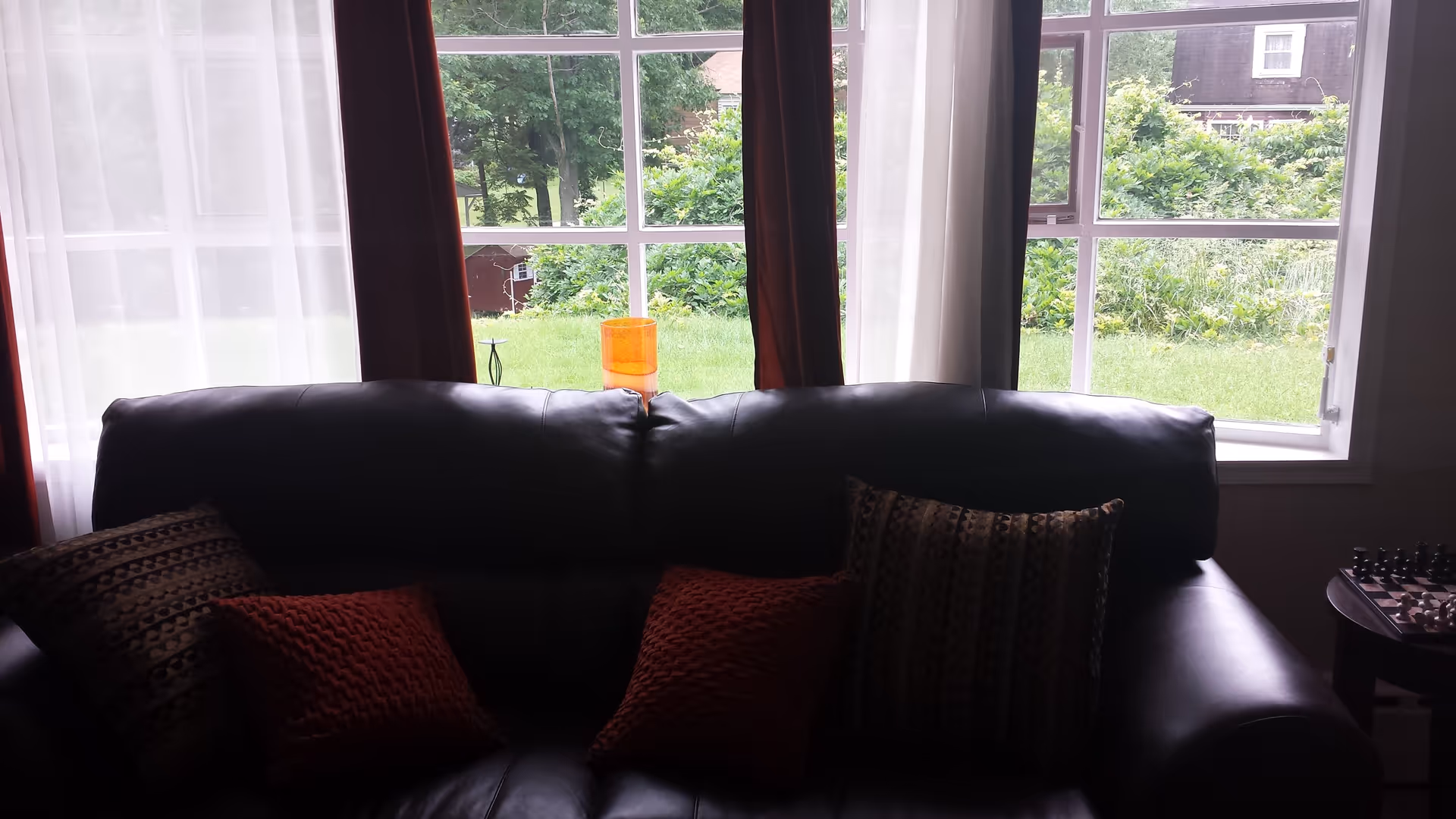 A dark leather couch with four decorative pillows in front of a large window with white and dark red curtains. Outside the window, there is a green lawn and trees. A small table with a chessboard is visible to the right of the couch.