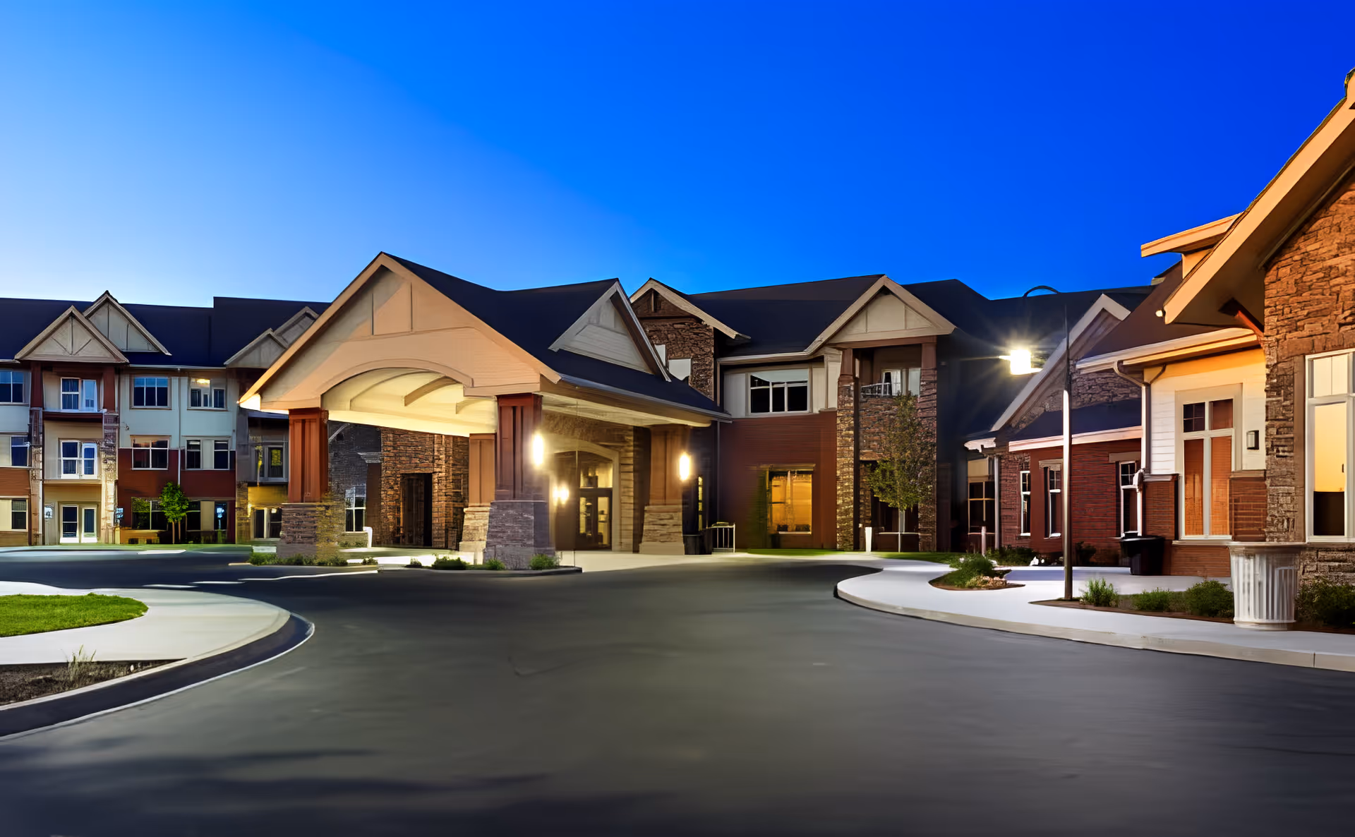 Exterior view of The Terraces of Boise senior living facility at dusk, showing a large covered entrance with stone and wood architectural details, well-lit windows, and a curved driveway with landscaped areas.