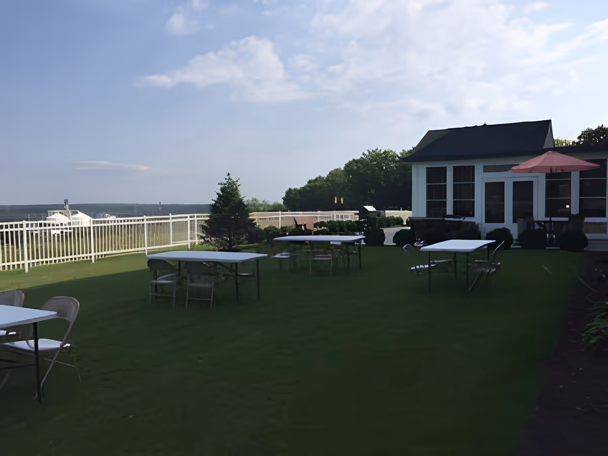 Outdoor patio area with several white tables and chairs on a green lawn. There is a white fence along the edge, a small tree, and a building with large windows and a red umbrella on the right side. The sky is partly cloudy.