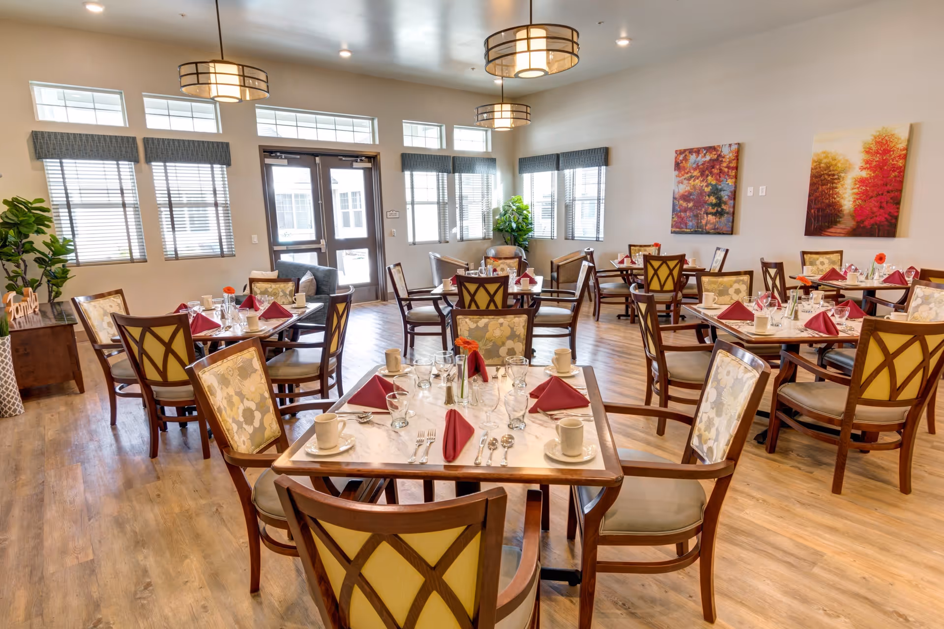 A bright dining room with multiple tables set for a meal, each table featuring maroon folded napkins, cups, glasses, and silverware. The room has wooden floors, large windows with blinds, modern ceiling lights, and colorful autumn-themed paintings on the walls.