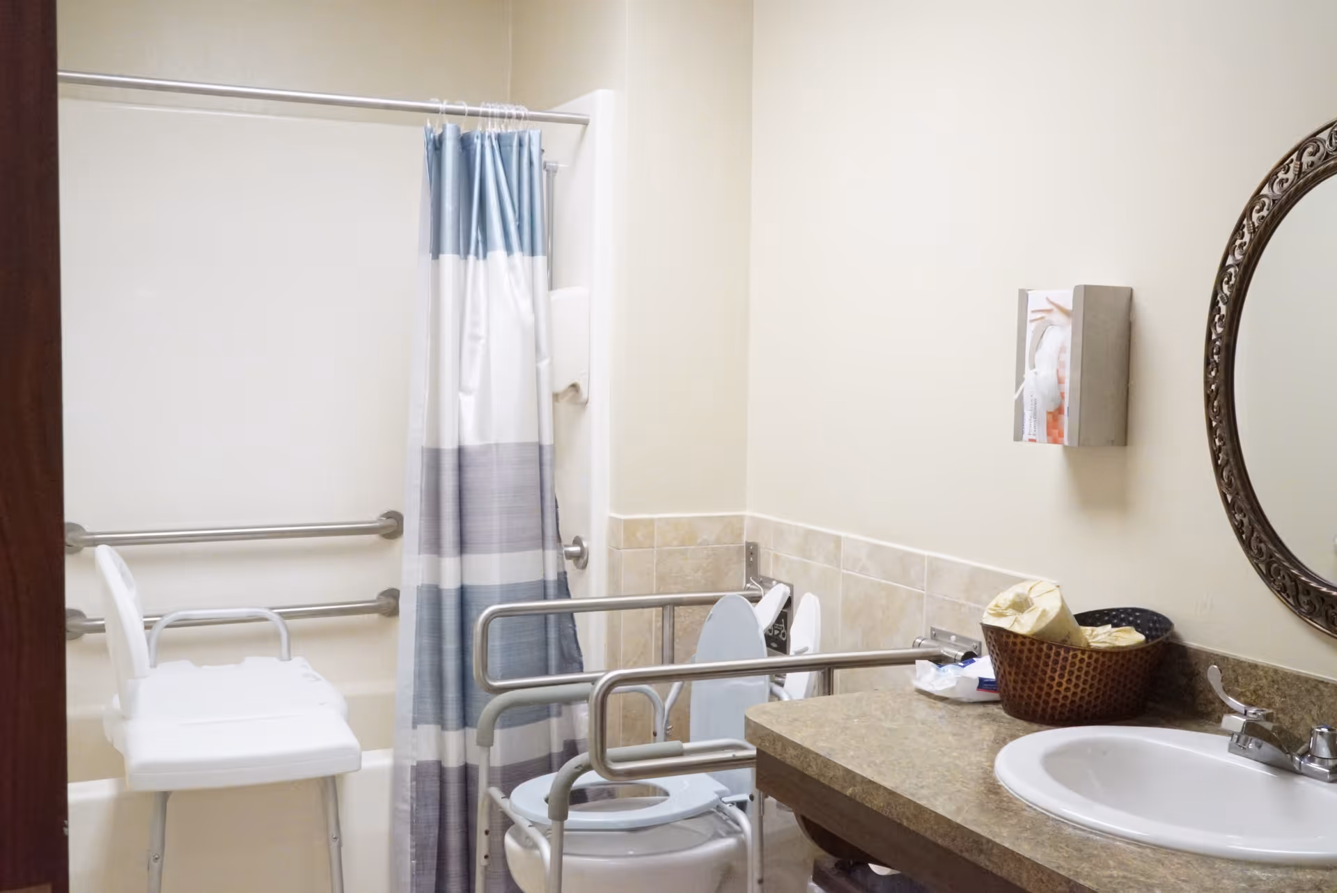 Accessible bathroom with a shower area featuring a white shower chair and grab bars, a toilet with safety rails, a countertop with a sink, a round decorative mirror, and a wall-mounted tissue dispenser.