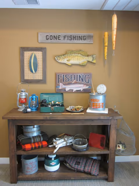 A rustic wooden table against a tan wall decorated with fishing-themed items including a wooden sign that says 'GONE FISHING', a mounted fish, a framed fishing lure, and a sign that says 'FISHING'. The table holds various fishing gear such as lanterns, a tackle box, a metal bucket, and other fishing accessories. Fishing floats hang from the ceiling on the right side.