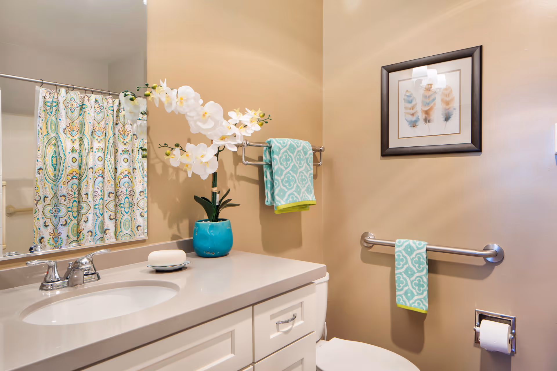Small beige bathroom featuring a sink vanity with a turquoise orchid, patterned shower curtain, and wall-mounted grab bars.