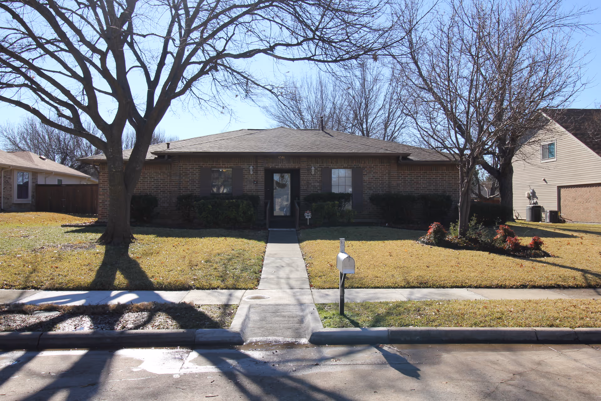 Single-story brick house with a front lawn, sidewalk, mailbox, and leafless trees.