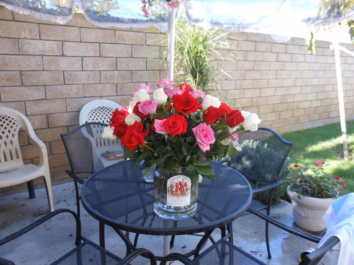 Outdoor patio area with a round black metal table holding a glass vase filled with red, pink, and white roses. Surrounding the table are several chairs, including black metal and white plastic ones. A brick wall and some green plants are visible in the background under a clear sky.