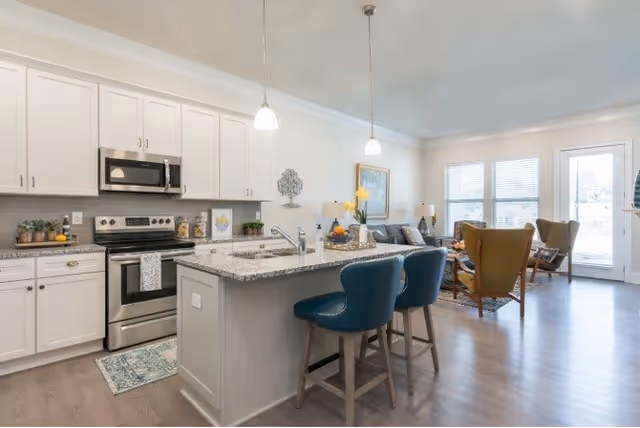 Bright and modern open-concept kitchen and living room area in a senior living facility. The kitchen features white cabinets, stainless steel appliances including a microwave and stove, a granite countertop island with a sink, and two blue bar stools. The living room has comfortable seating with armchairs and a sofa, a coffee table, and large windows letting in natural light.