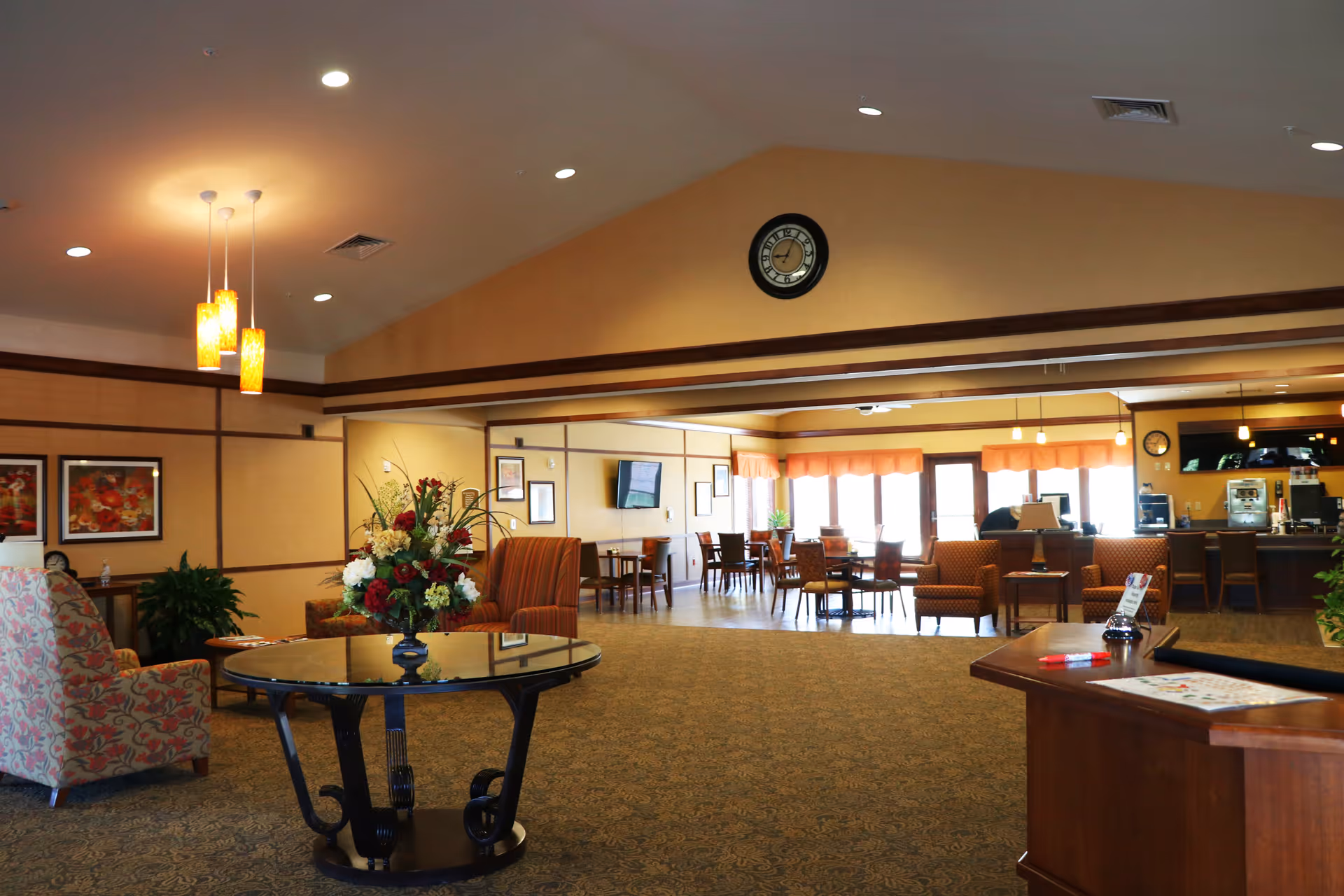Spacious and well-lit common area in a senior living facility featuring comfortable armchairs, a round glass table with a floral arrangement, multiple dining tables and chairs near large windows with orange valances, a wall clock, and a counter area with coffee machines and bar stools.