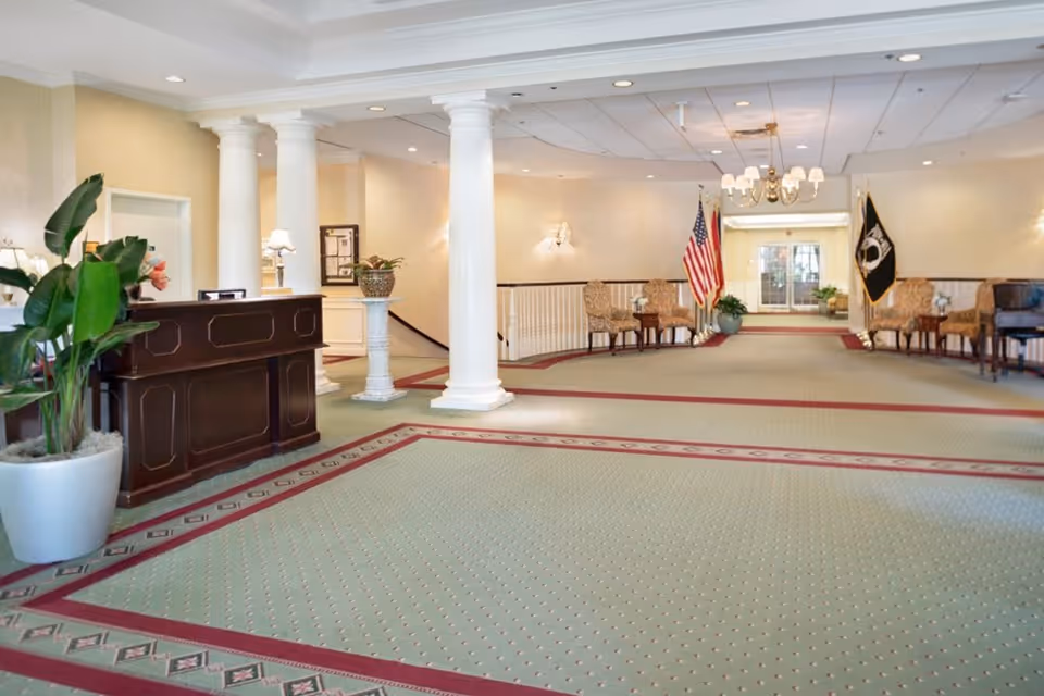 Spacious lobby area with green carpet bordered in red, white columns, a dark wooden reception desk, potted plants, American and POW flags, and several upholstered chairs arranged along the walls under soft lighting.