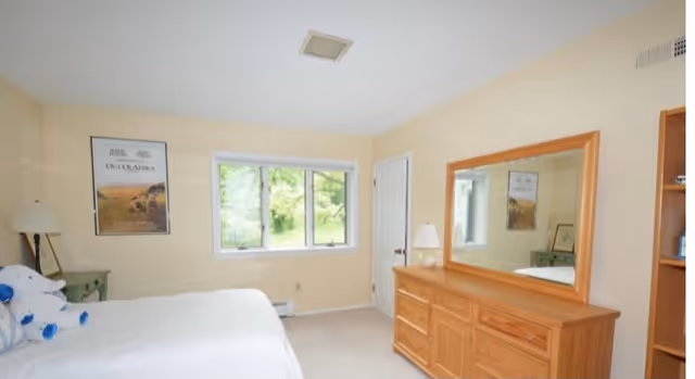 A bedroom with a large window showing greenery outside, a bed with white bedding and a blue and white stuffed animal, a green nightstand with a lamp, a wooden dresser with a large mirror above it, and a door next to the window.