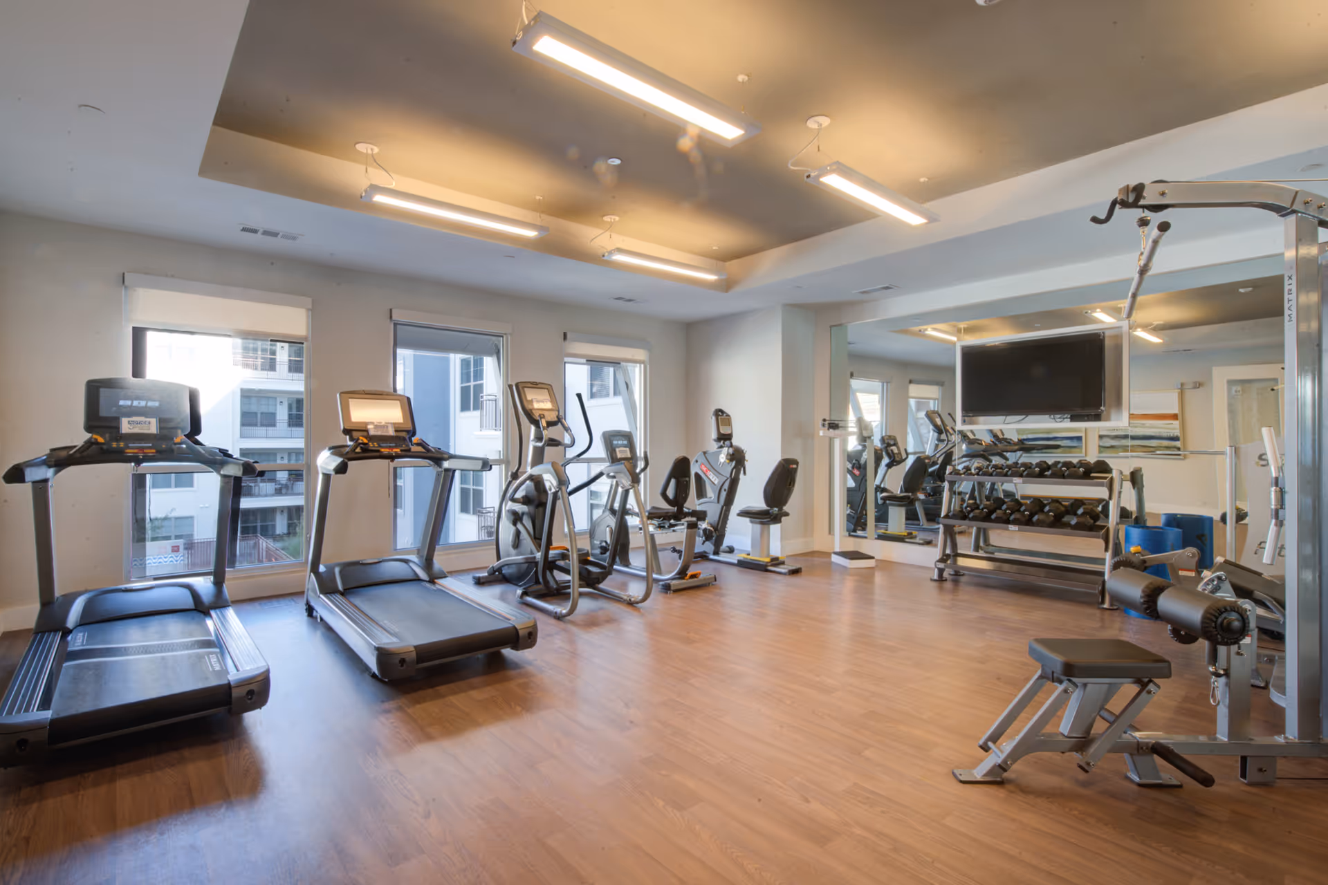 A modern fitness room with wooden flooring, two treadmills, two elliptical machines, a recumbent bike, a weight bench, a rack of dumbbells, and a mounted flat-screen TV. Large windows allow natural light to enter the room, and a large mirror covers one wall.
