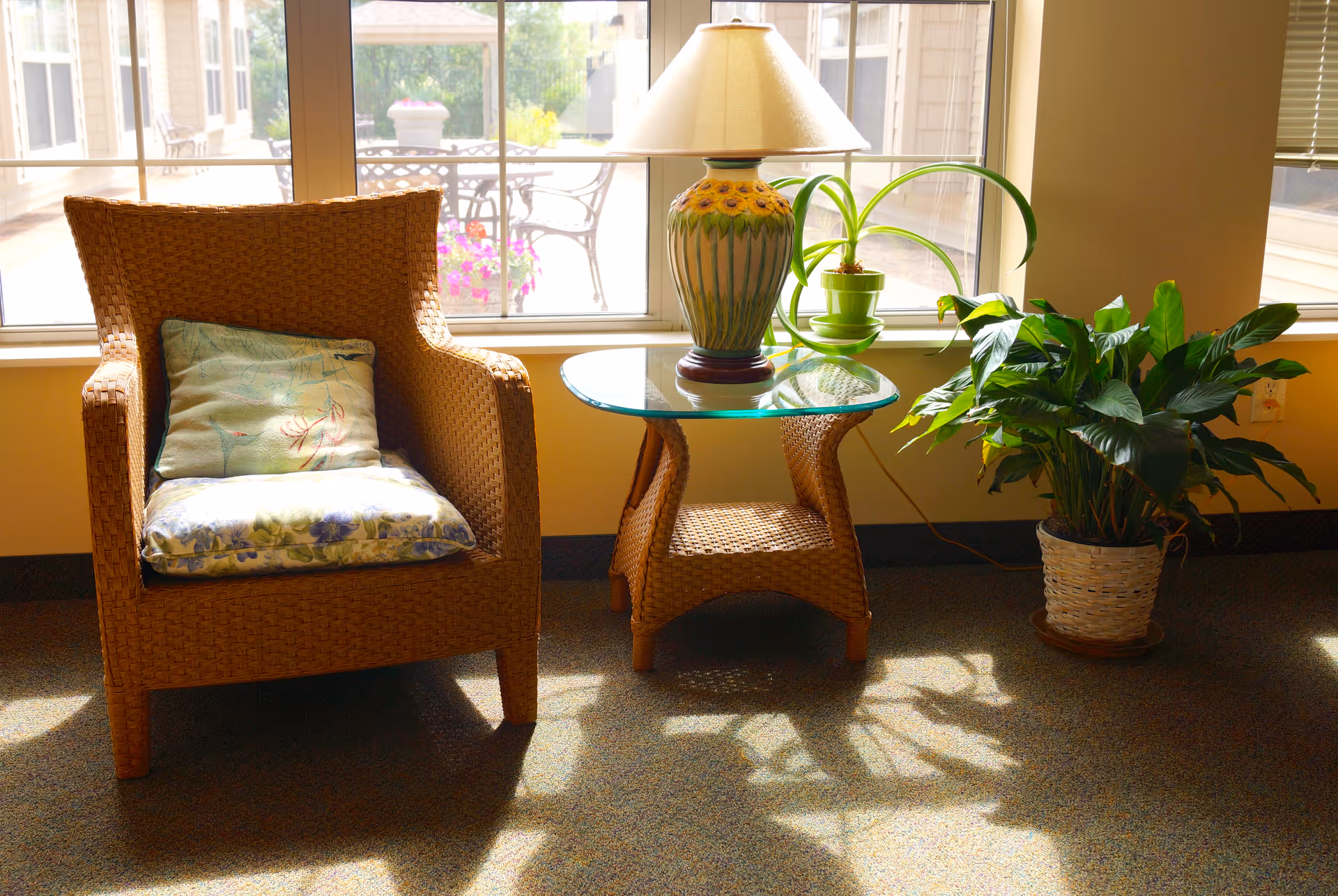 A cozy corner with a wicker armchair featuring floral cushions, a glass-top wicker side table with a decorative lamp and a small potted plant, and a larger potted plant on the floor next to the table. Sunlight streams through large windows behind the furniture, illuminating the space.