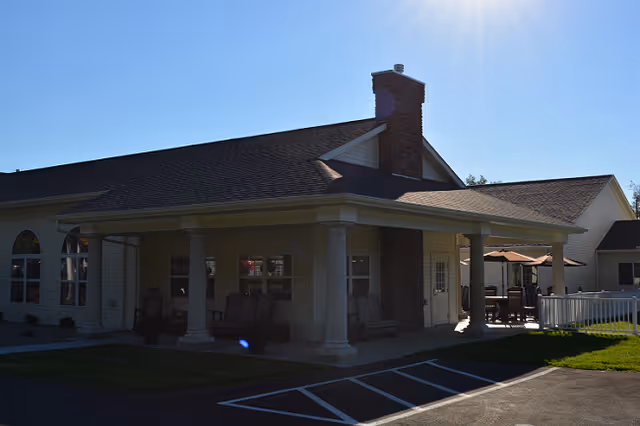 Single-story assisted living building with a covered porch supported by columns, outdoor seating, and a chimney under a bright sunny sky.