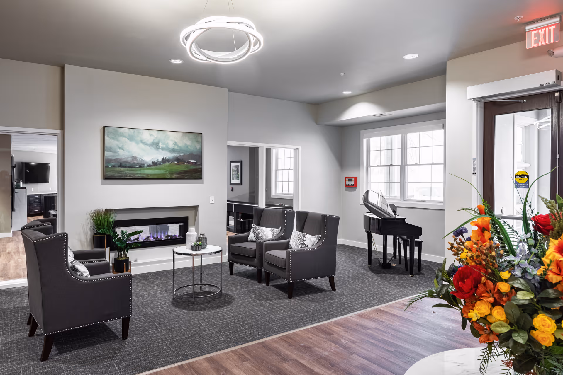 Bright modern senior living lobby with upholstered seating around a fireplace, a piano by the window, and a large floral arrangement near the entrance.