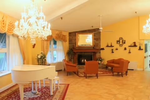 Spacious elegant lounge with a white grand piano, crystal chandeliers, a seating area around a stone fireplace, and yellow walls.