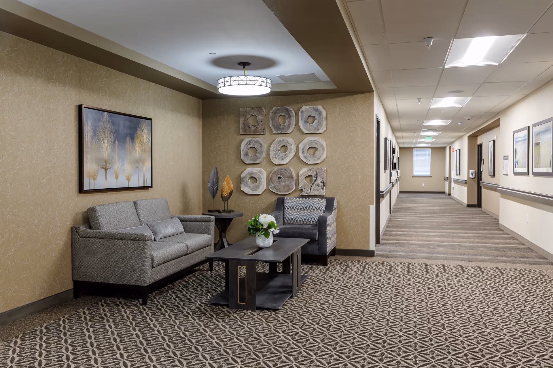 A furnished seating area with a sofa, armchair, coffee table, and decorative wall art beside a long carpeted hallway.