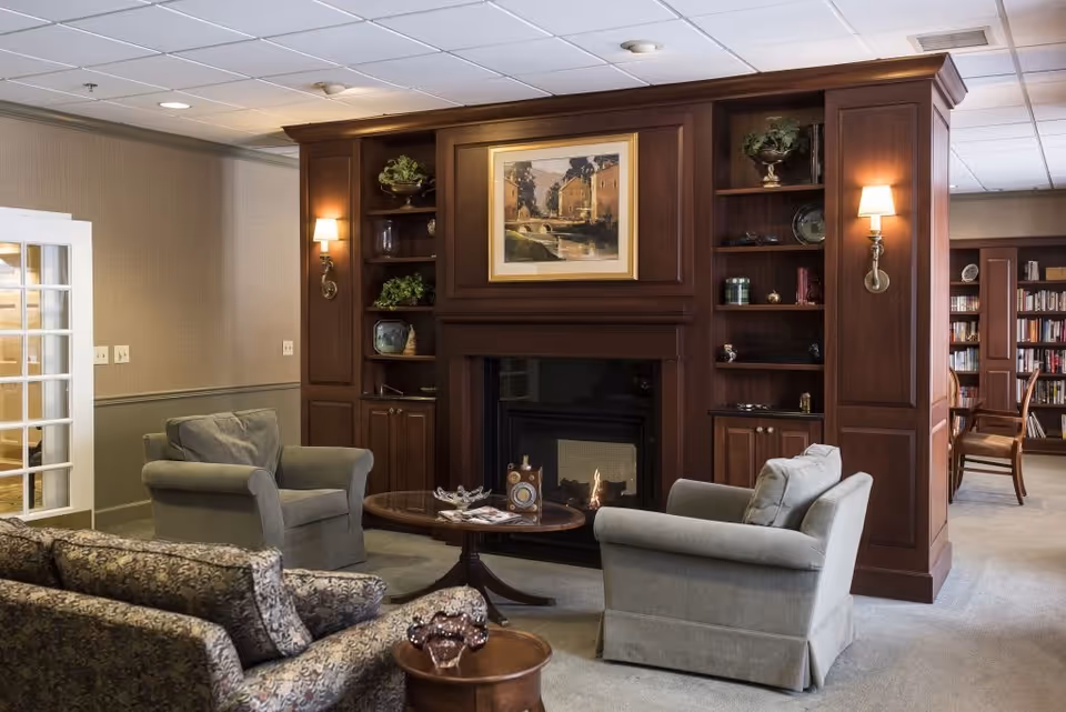 A cozy living room area in a senior living facility featuring a wooden fireplace with built-in shelves on either side, decorated with plants and ornaments. Two gray armchairs and a patterned sofa are arranged around a round wooden coffee table with magazines and decorative items. Wall sconces provide warm lighting, and a framed painting hangs above the fireplace. In the background, a bookshelf and additional seating are visible.