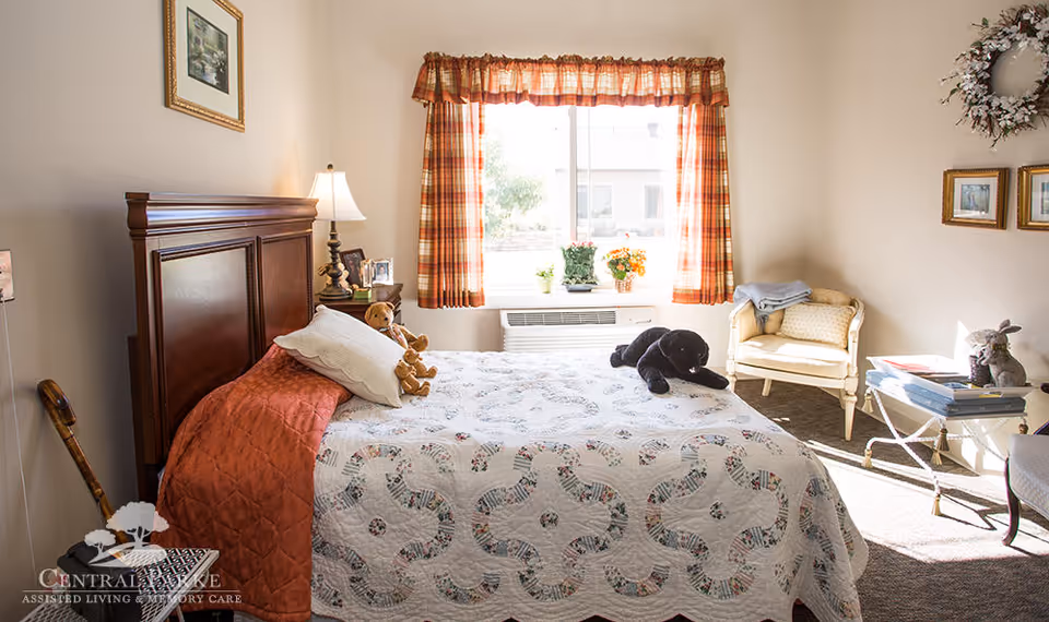 Bright, traditionally furnished bedroom with a made bed, window with plaid curtains, and an armchair.