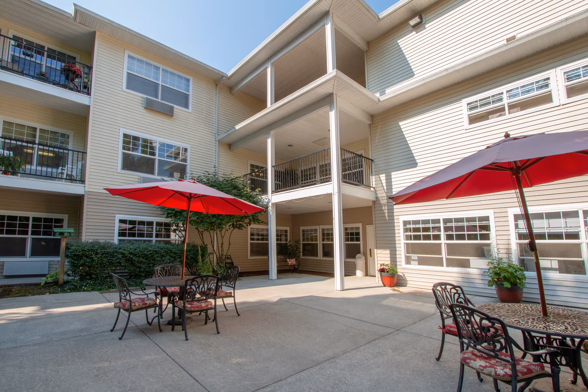 Outdoor courtyard area of a senior living facility with beige siding buildings surrounding a concrete patio. The patio has two tables with red umbrellas and black metal chairs with patterned cushions. There are potted plants near the building walls and balconies with railings on the upper floors.