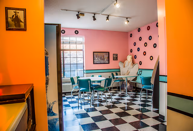 Retro-style dining room with black-and-white checkered floor, turquoise diner chairs and round tables, pink walls decorated with records and framed art.