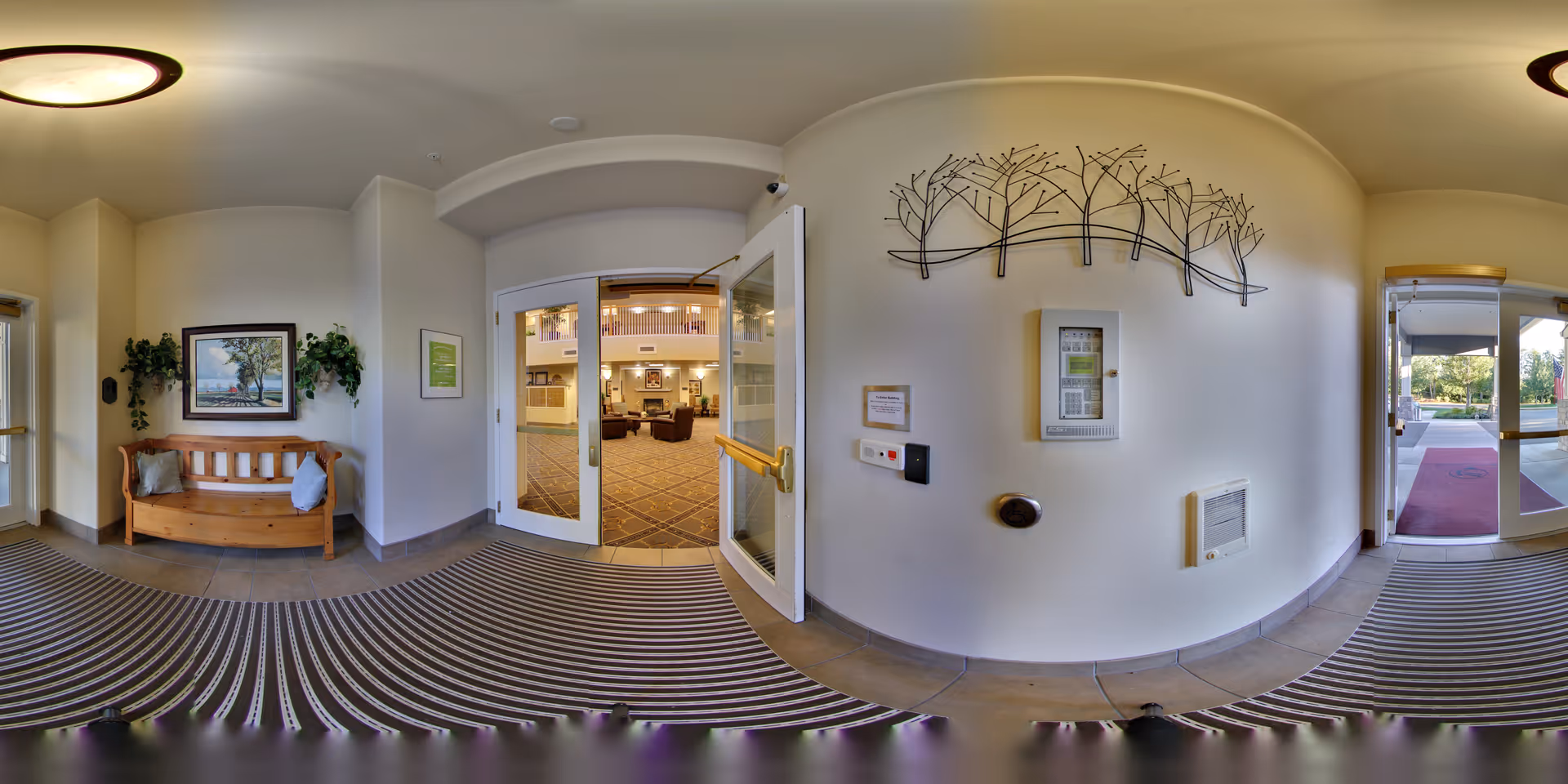 Entrance vestibule with a wooden bench, double glass doors opening into a lobby, and decorative wall art on the curved wall.