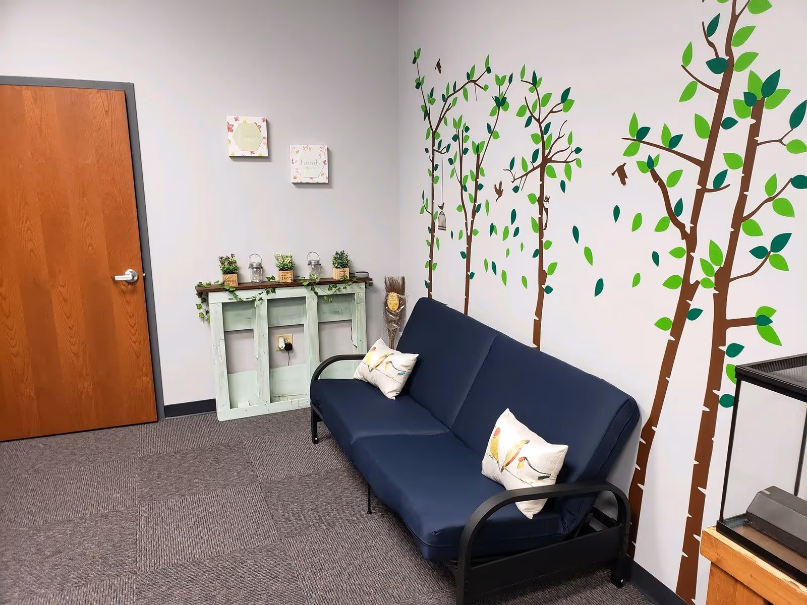 A small room with a blue couch featuring two bird-themed pillows, a wall decorated with tree and bird decals, a wooden door, and a small rustic table with potted plants and decorative items.