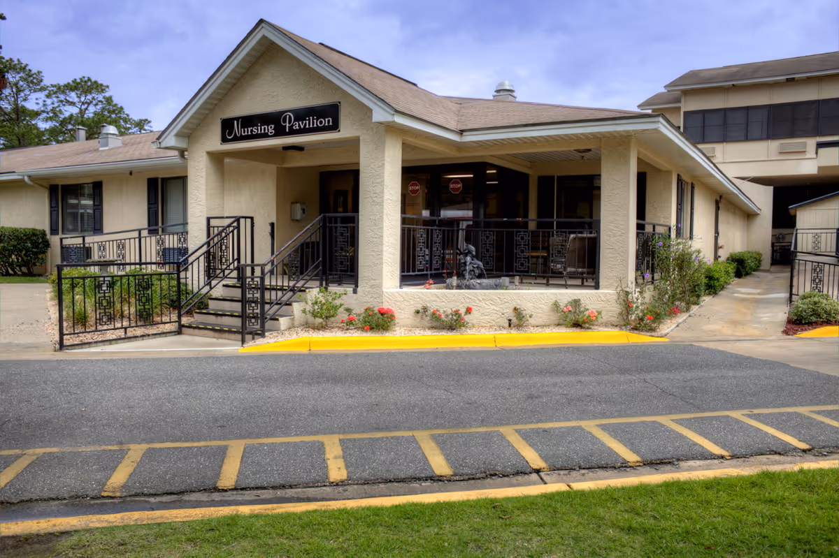 Exterior view of the Nursing Pavilion at Chipola Health and Rehabilitation Center, showing a beige building with a ramp and stairs leading to the entrance, black railings, a small garden with flowers, and a paved driveway with yellow markings.