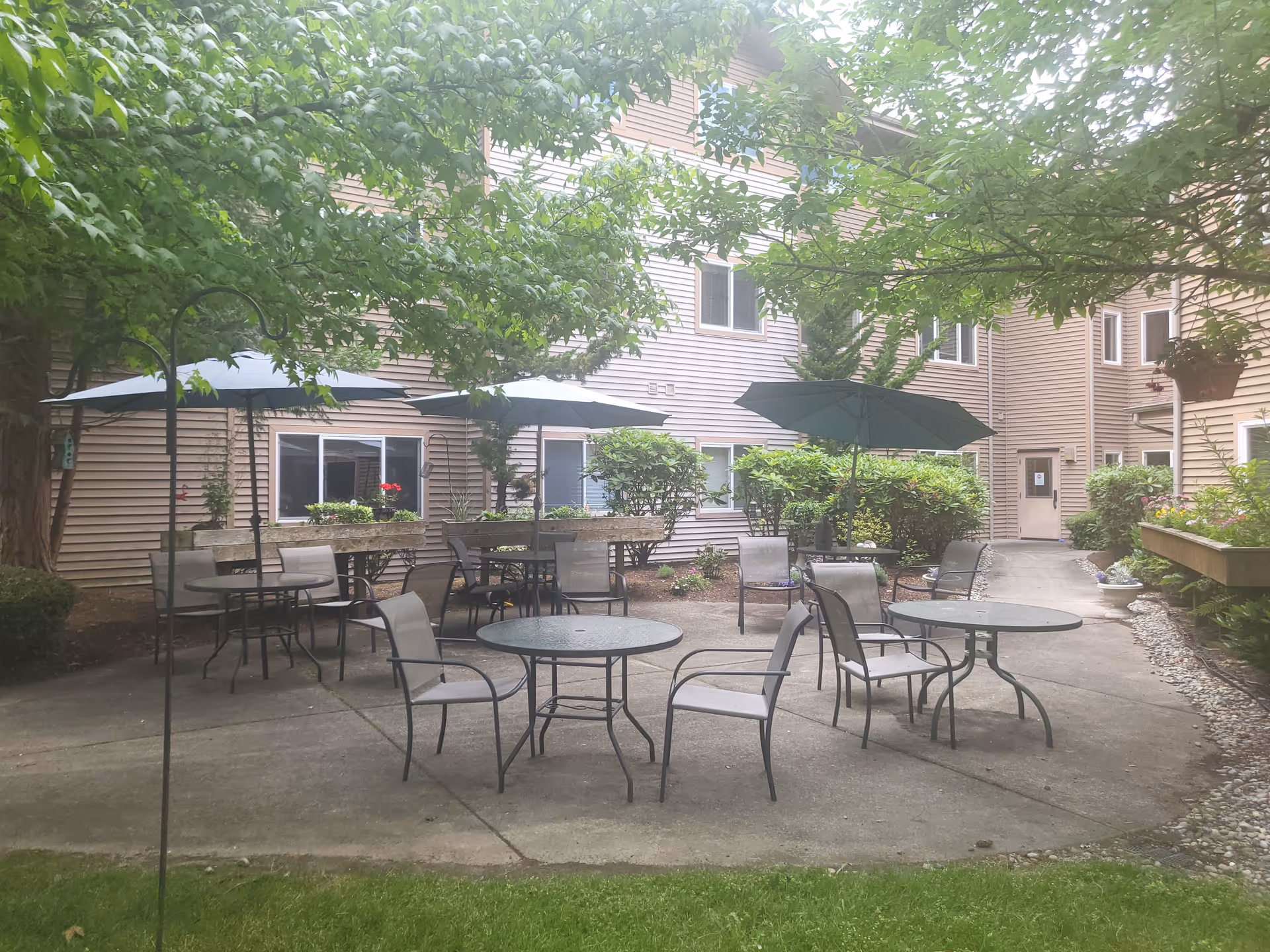 Outdoor patio area at Vineyard Park of Port Orchard with several round metal tables and chairs, some shaded by large umbrellas. The patio is surrounded by greenery including bushes and trees, with the building's beige siding and windows visible in the background.