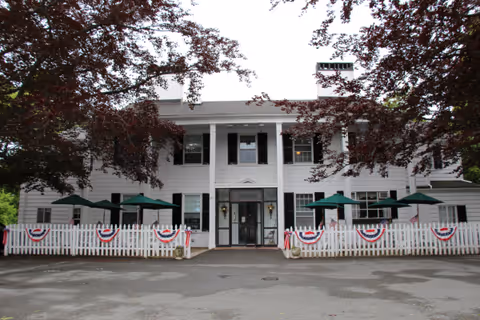 Front exterior view of Jesmond Nursing Home, a two-story white building with black shutters, a white picket fence decorated with red, white, and blue bunting, and green patio umbrellas on the porch. Trees with dark red leaves frame the building.