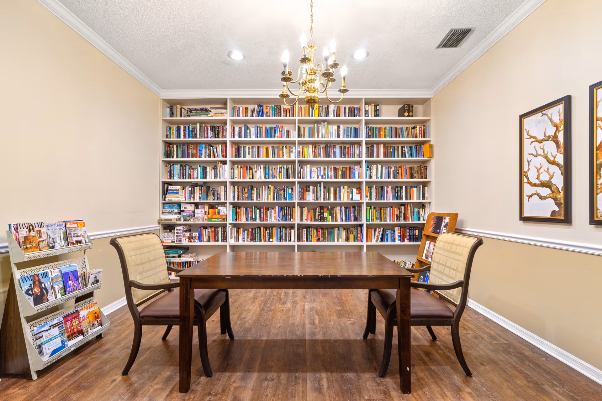 A quiet reading room with a large wooden table and two cushioned chairs facing a wall filled with bookshelves. The shelves are stocked with numerous books. To the left, there is a rack holding magazines. The room has wooden flooring, beige walls with white trim, two framed tree artwork pieces on the right wall, and a brass chandelier hanging from the ceiling.