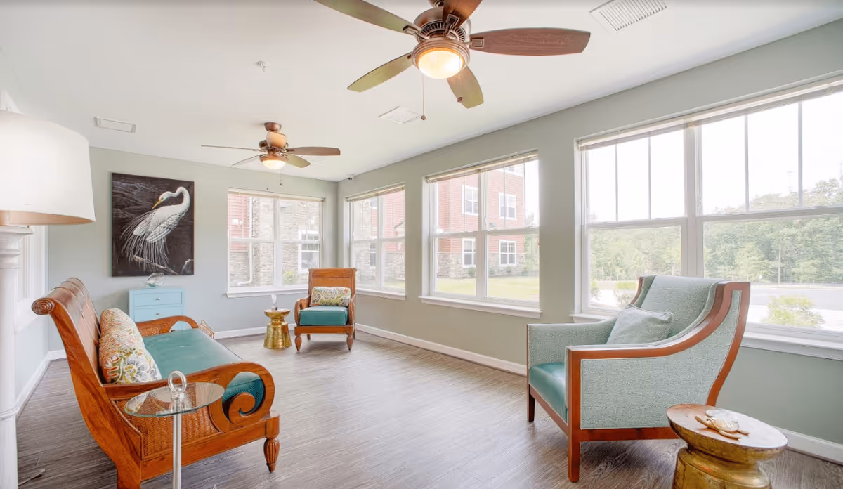 A bright and airy living room with large windows letting in natural light. The room features wooden framed seating with teal cushions, a small round side table, ceiling fans with lights, and a painting of a white bird on the wall. The floor is wood-style and the walls are painted light green.