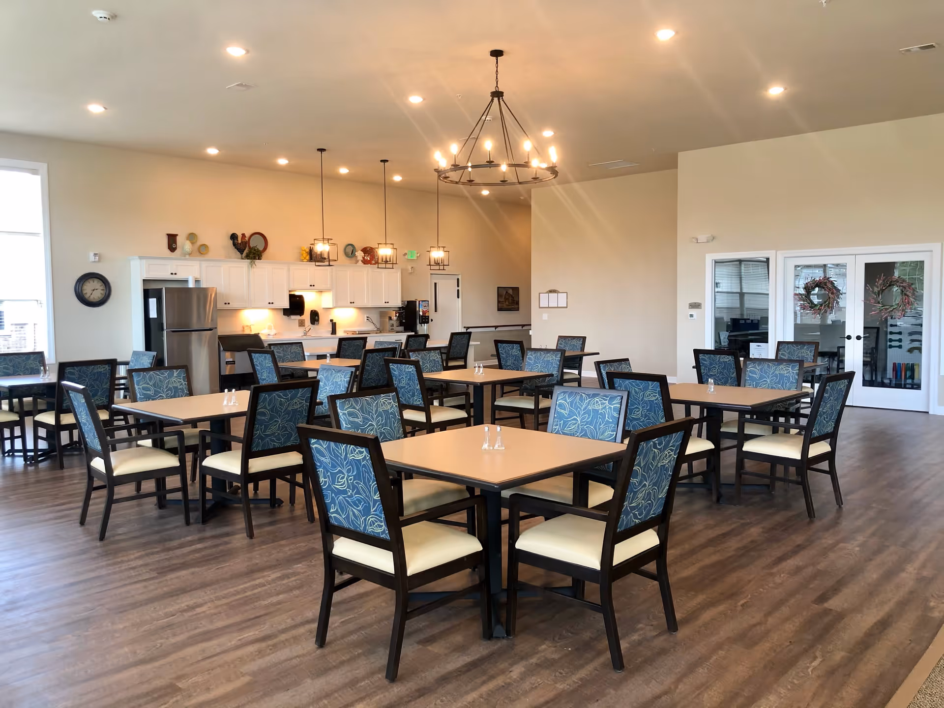 Bright dining area with multiple square tables and upholstered chairs and a small kitchen/service counter in the background.